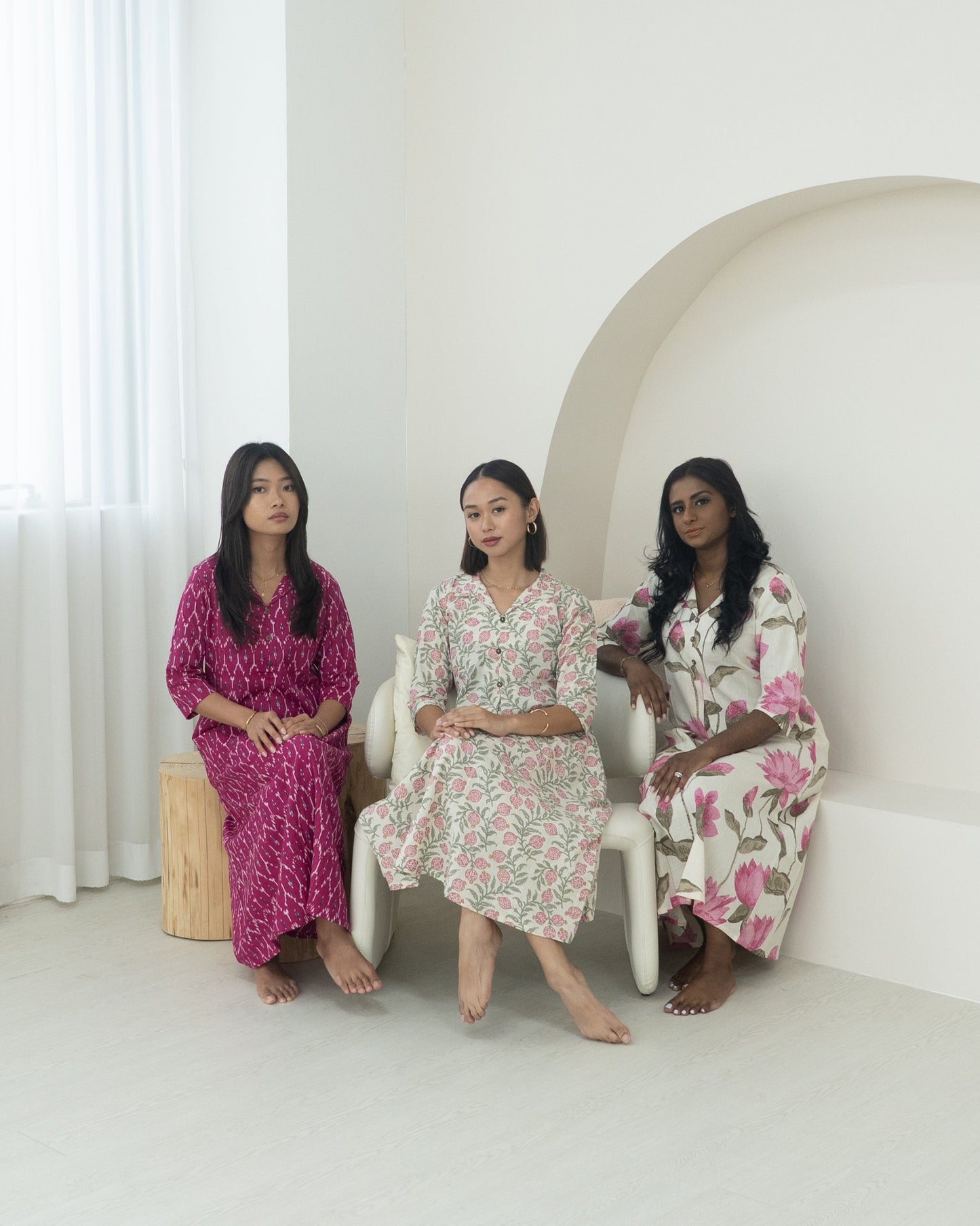 Three women sitting on a white chair in a minimalistic room with a white wall and light-colored floor.