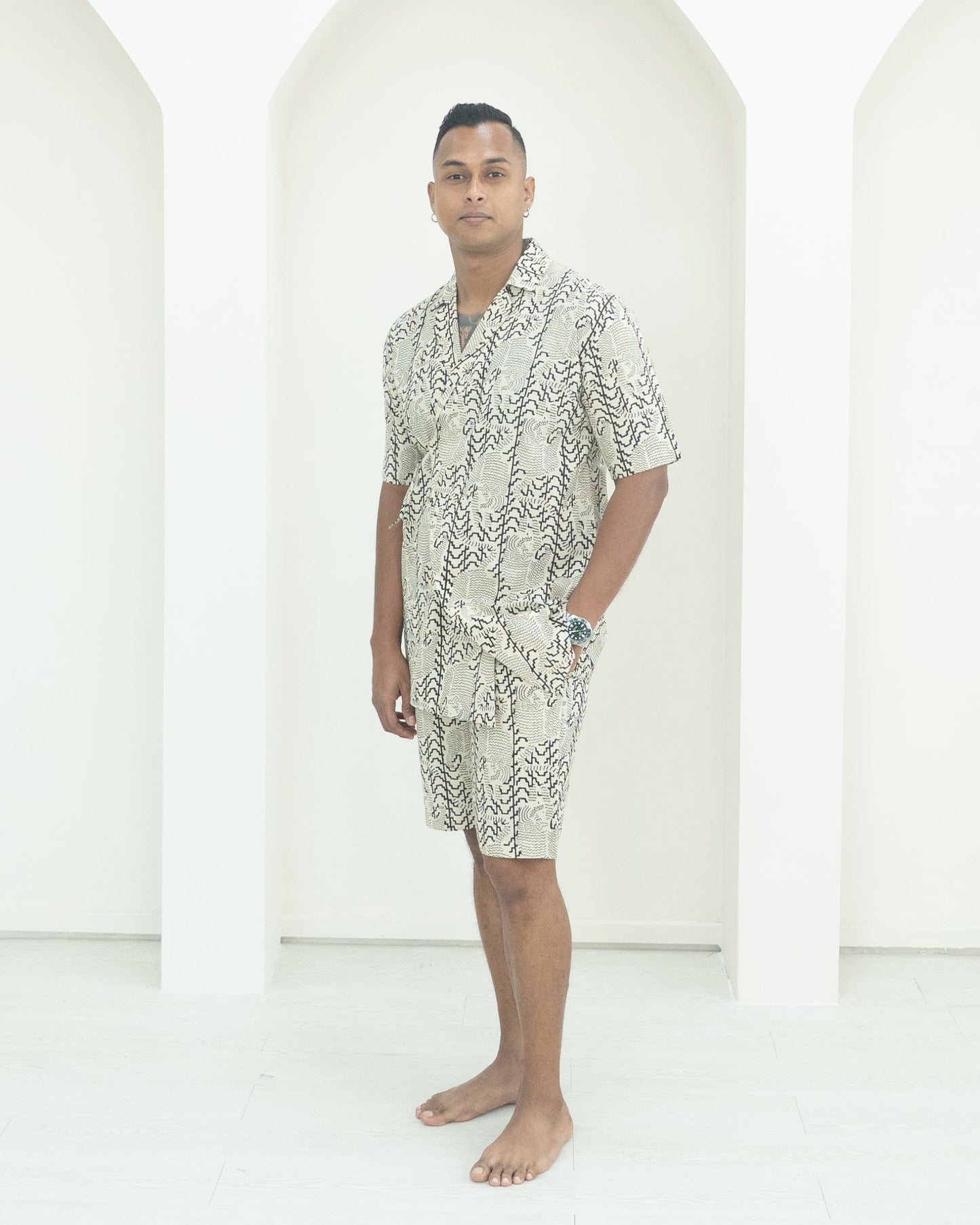 Man wearing a patterned shirt and shorts standing in front of white background.