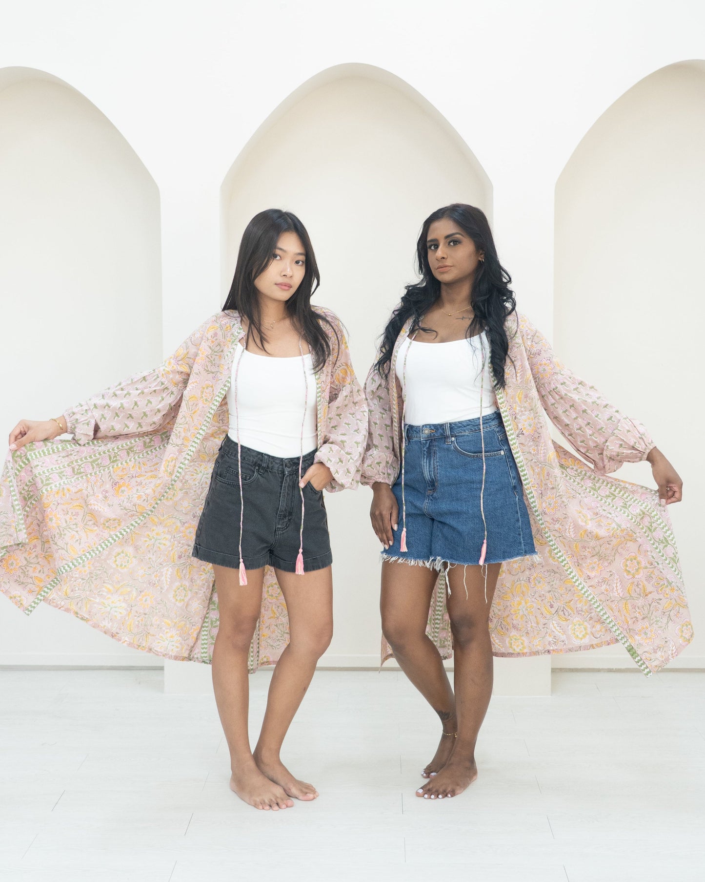 Two women standing side by side against a white wall wearing pink overcoats