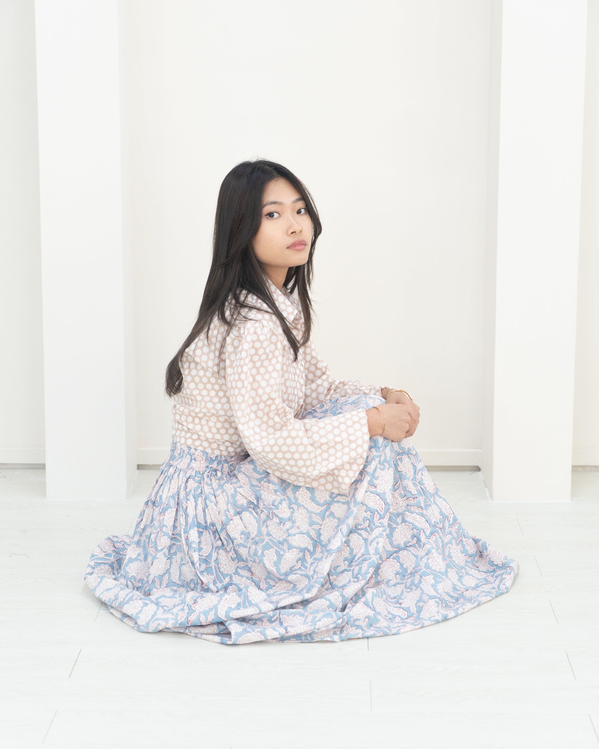 Woman sitting on a white floor wearing a white blouse with polka dots and a blue floral skirt.