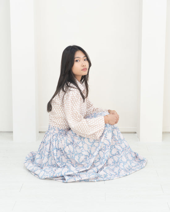 Woman sitting on a white floor wearing a white blouse with polka dots and a blue floral skirt.