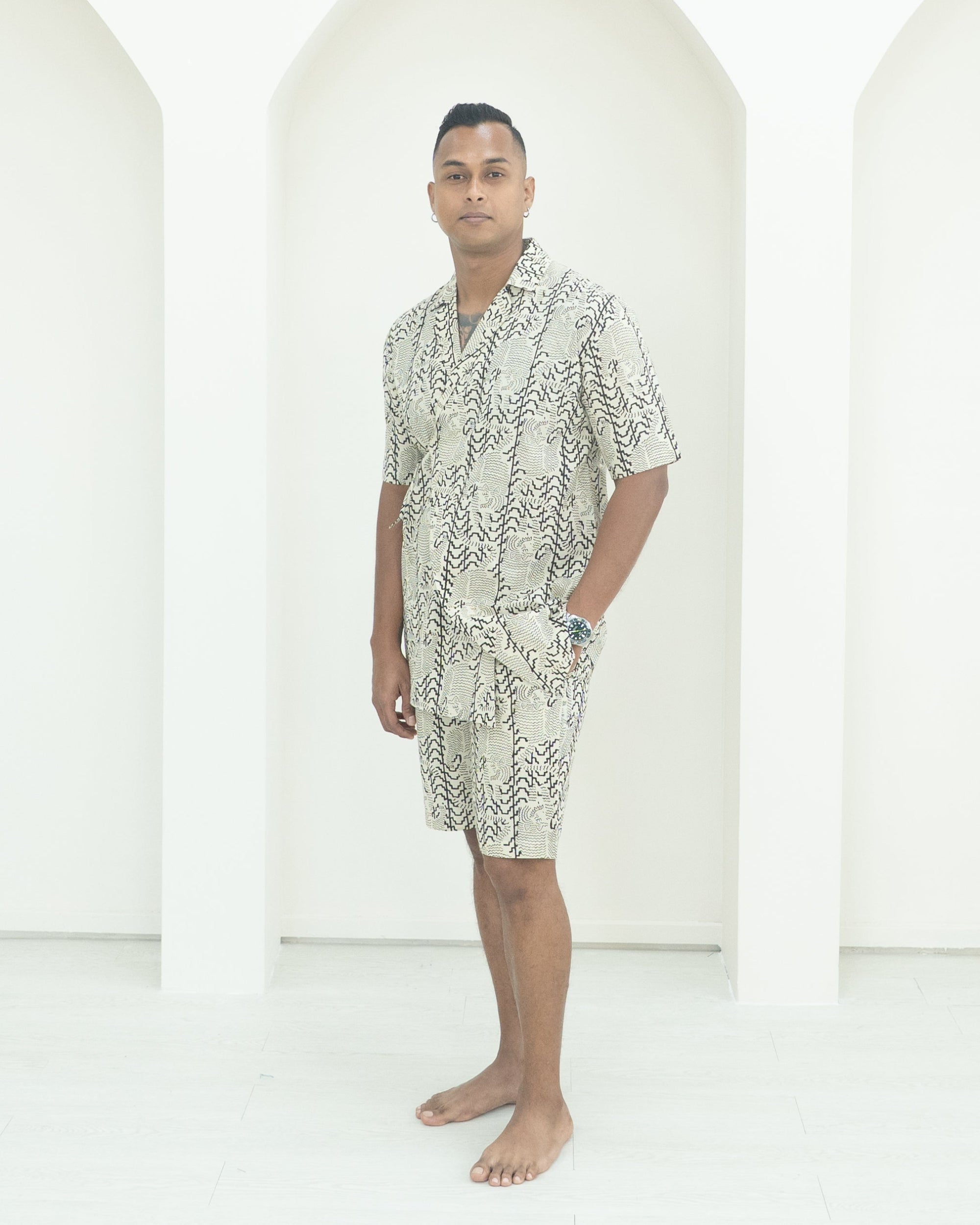 Man wearing a patterned shirt and shorts standing in front of white background.