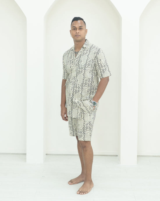 Man wearing a patterned shirt and shorts standing in front of white background.