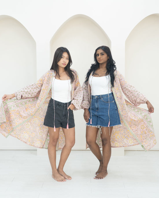 Two women standing side by side against a white wall wearing pink overcoats