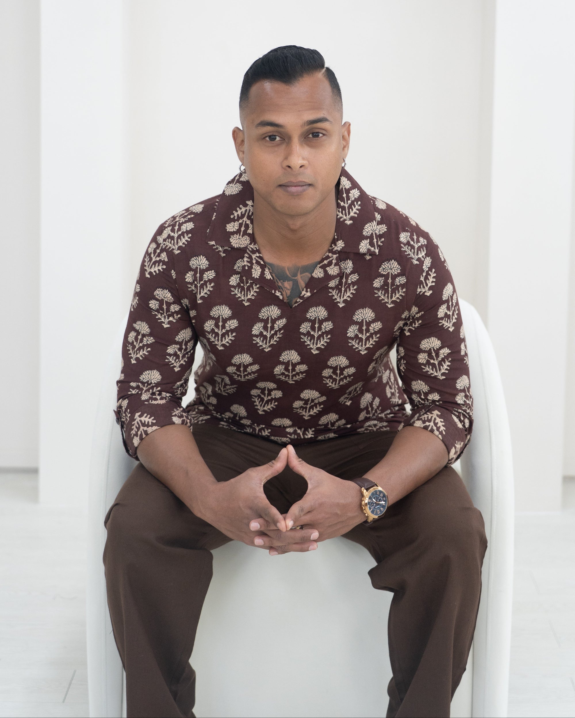 Man sitting on a white architectural structure wearing a patterned shirt and brown pants.