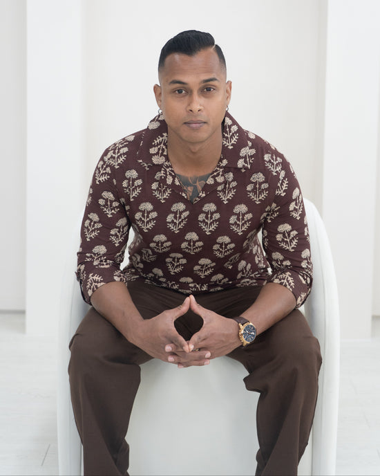 Man sitting on a white architectural structure wearing a patterned shirt and brown pants.